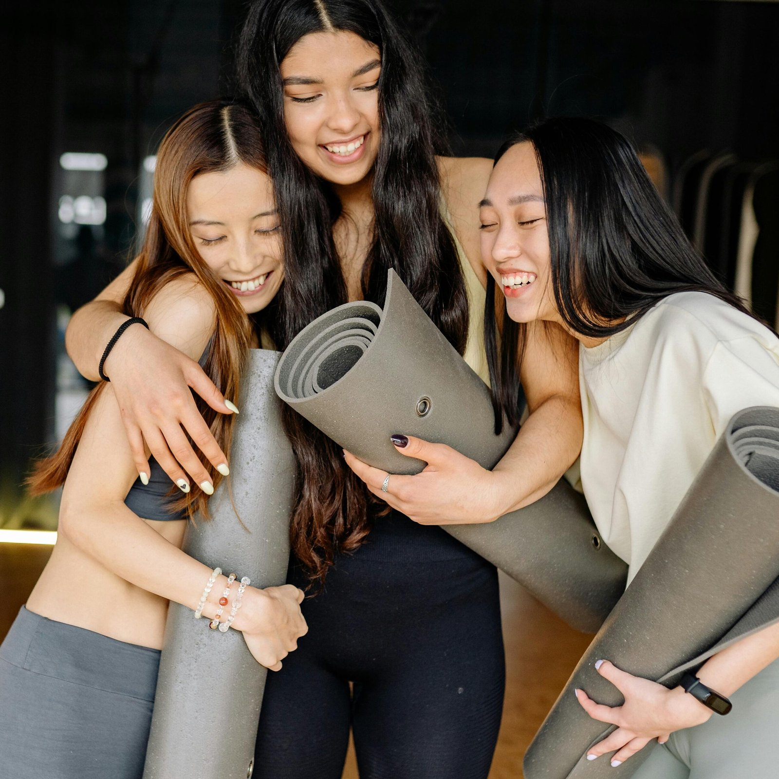 Three young women joyfully hugging and holding yoga mats, symbolizing friendship and healthy living.